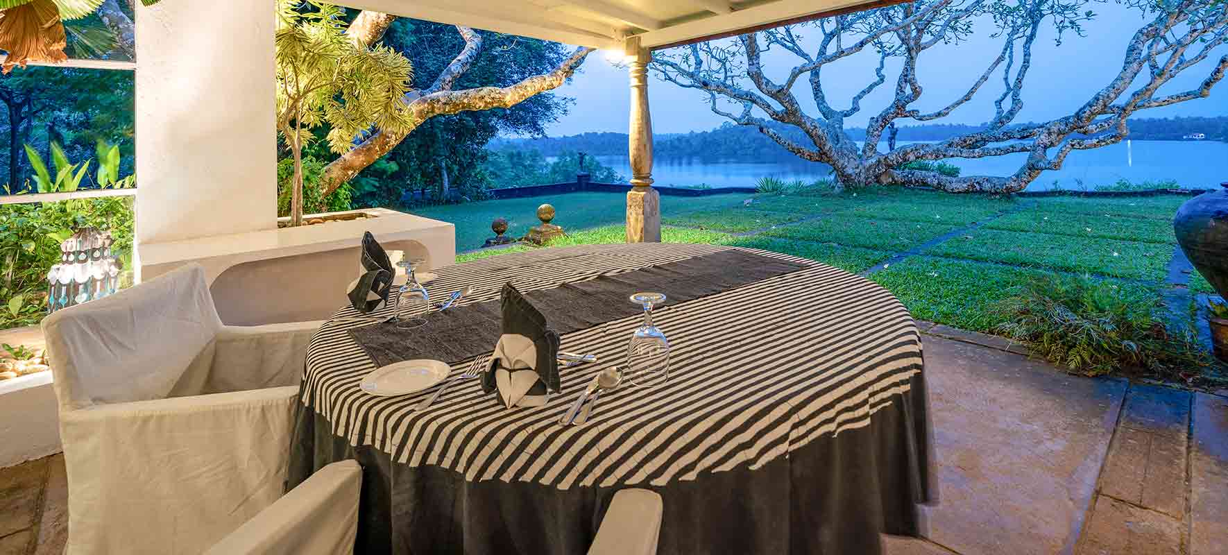 A table laid on a covered veranda, looking across a lawn and a frangipani tree to the lake
